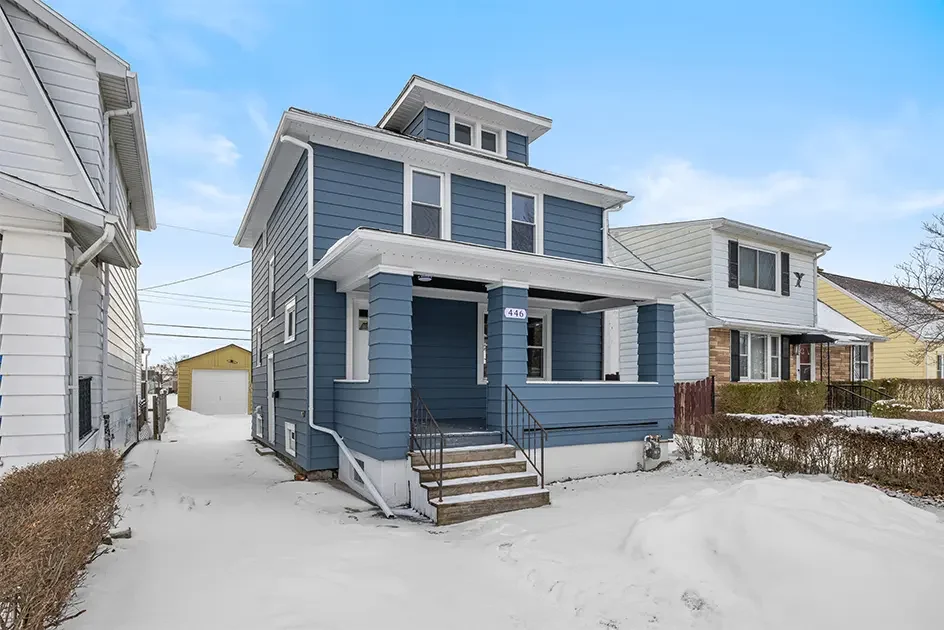 Exterior winter photo of 446 Victoria in Kenmore, NY, a blue two-story house with a covered front porch, snow-covered yard, and detached garage.