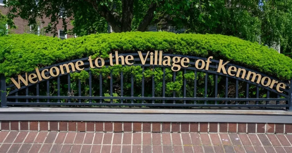 Welcome to the Village of Kenmore sign mounted on a black metal fence with green shrubs behind it.