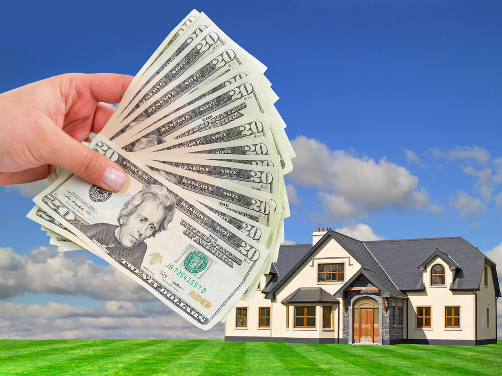 A hand holds cash in front of a beautiful home. Finding the best home buyer company is an important choice to sell your house fast in New Orleans.