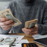 A person counting money on a table with a laptop next to them