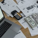 A table with a laptop, calculator, papers, and cash