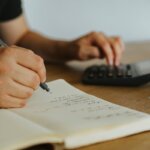 A person typing on a calculator and writing stuff down on paper