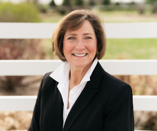 Melanie Jones Realtor in a Black Blazer in front of a white board fence.