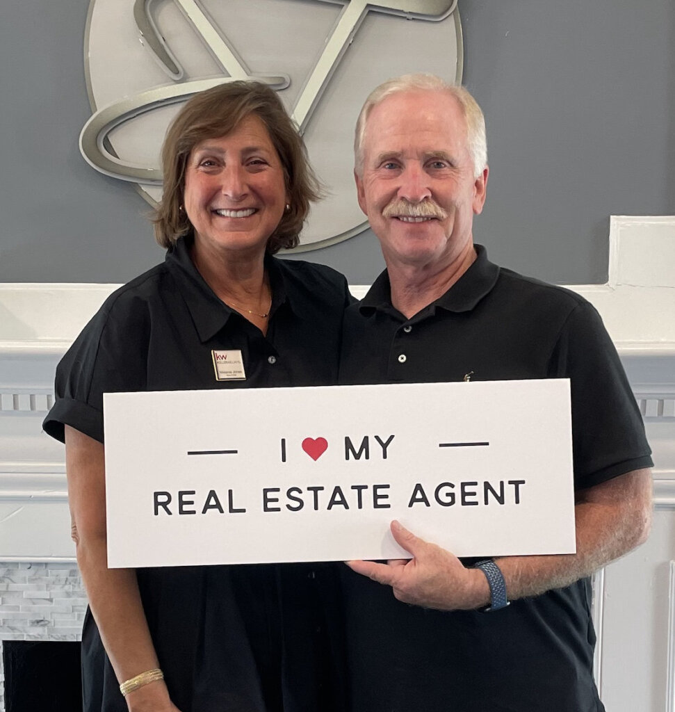 John and Melanie holding an "I love My Real Estate Agent" sign at a closing in front of a white fireplace mantel with grey walls.