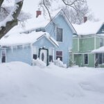 A blue and white house with a winter-ready roof is covered in snow.