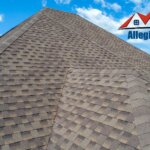 A man is standing on top of a shingled roof, inspecting a clogged gutter.