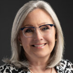 Becky Fields the Grandma Who Buys Houses headshot in her b/w jacket, glasses, and smiling, shoulder length silver gray hair.