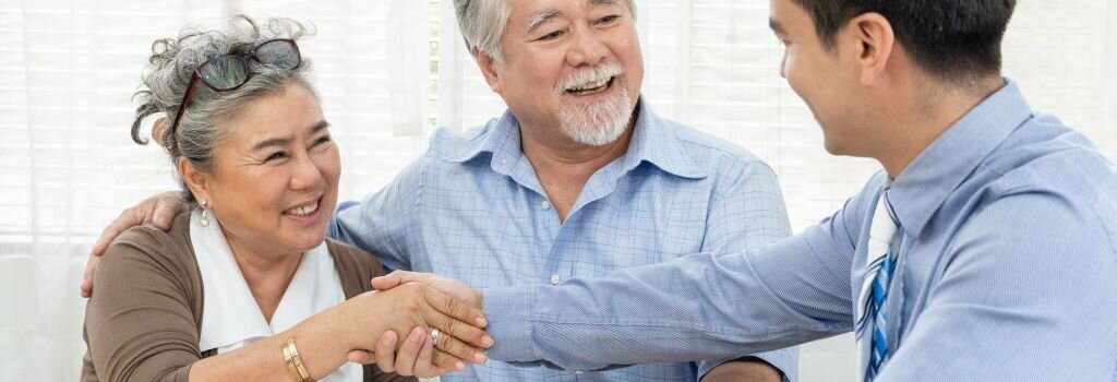 An older homeowner couple shaking hands with an advisor at a table, completing a straightforward home sale decision with calm confidence