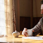 A man sits alone at a kitchen table holding a cup of coffee, looking out the window, with papers and an alarm clock resting on the table nearby.