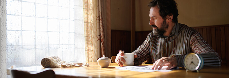 A man sits alone at a kitchen table holding a cup of coffee, looking out the window, with papers and an alarm clock resting on the table nearby.