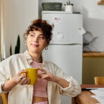 A young woman relaxes with a yellow mug in her cozy kitchen at the table with papers looking out the window
