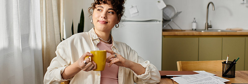 A young woman relaxes with a yellow mug in her cozy kitchen at the table with papers looking out the window with a confident smile