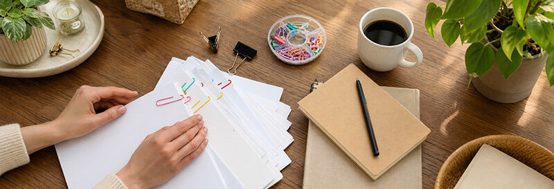 Hands sorting through paperwork and documents at a kitchen table with pencil, pad, and coffee