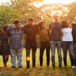 group of siblings and their significant others representing three generations as parr of a large family in a park with a setting sun behind them locked arms around their shoulders