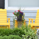 two yellow lawn chars sitting on the porch with a matching table and planter in between next to a blue house and rear window.