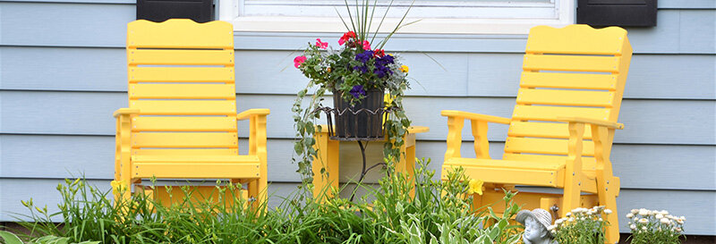 two yellow lawn chars sitting on the porch with a matching table and planter in between next to a blue house.