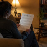 woman at home reading notice of sale foreclosure document sitting on a couch with a lamp lit room with toys strewn about on the floor