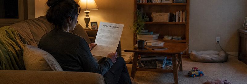 woman reading notice of sale foreclosure document sitting on a couch with a lamp lit room with toys strewn about on the floor
