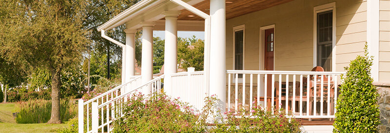 southern style front porch with yellow siding on a sunny day with four Greek columns and white picket railing conveying quiet care from a distance.