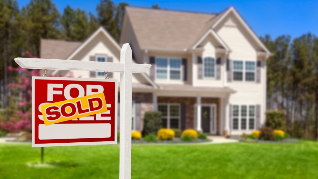 A suburban house with a 'For Sale' sign in the front yard marked with a 'Sold' label.