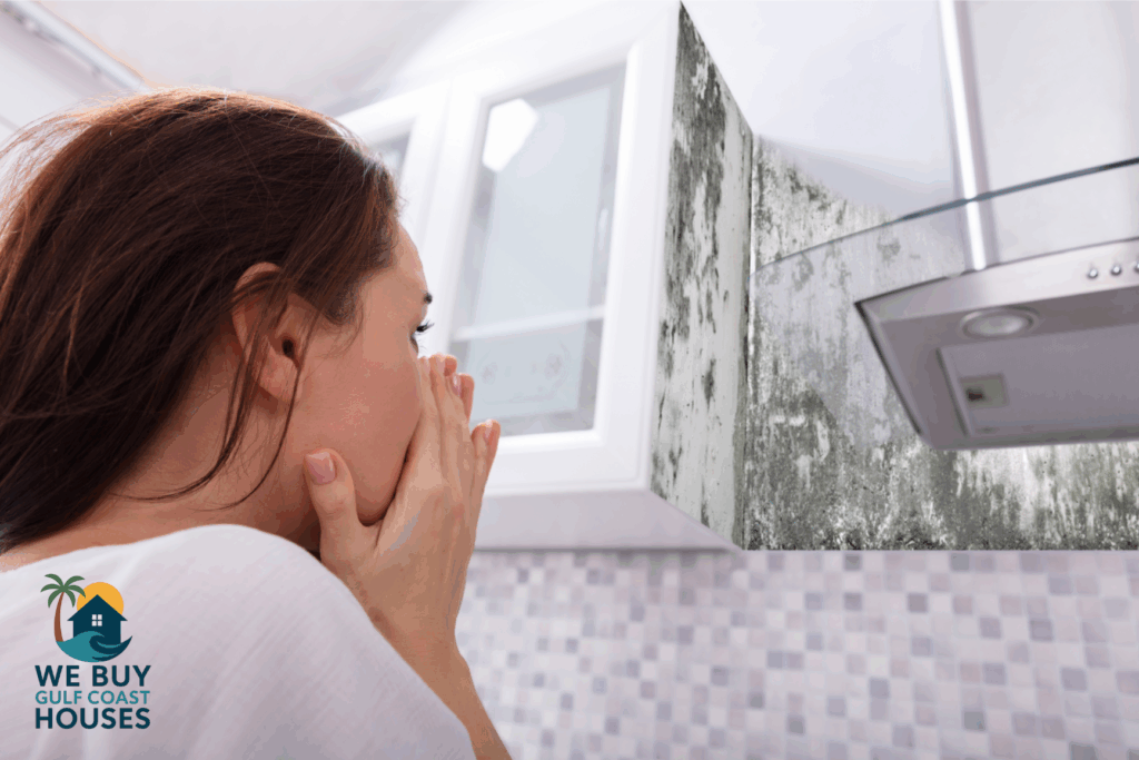 Woman in a modern kitchen reacts in shock to black mold growing up the wall behind the stove hood, showing a serious moisture issue.