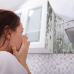 Woman in a modern kitchen reacts in shock to black mold growing up the wall behind the stove hood, showing a serious moisture issue.