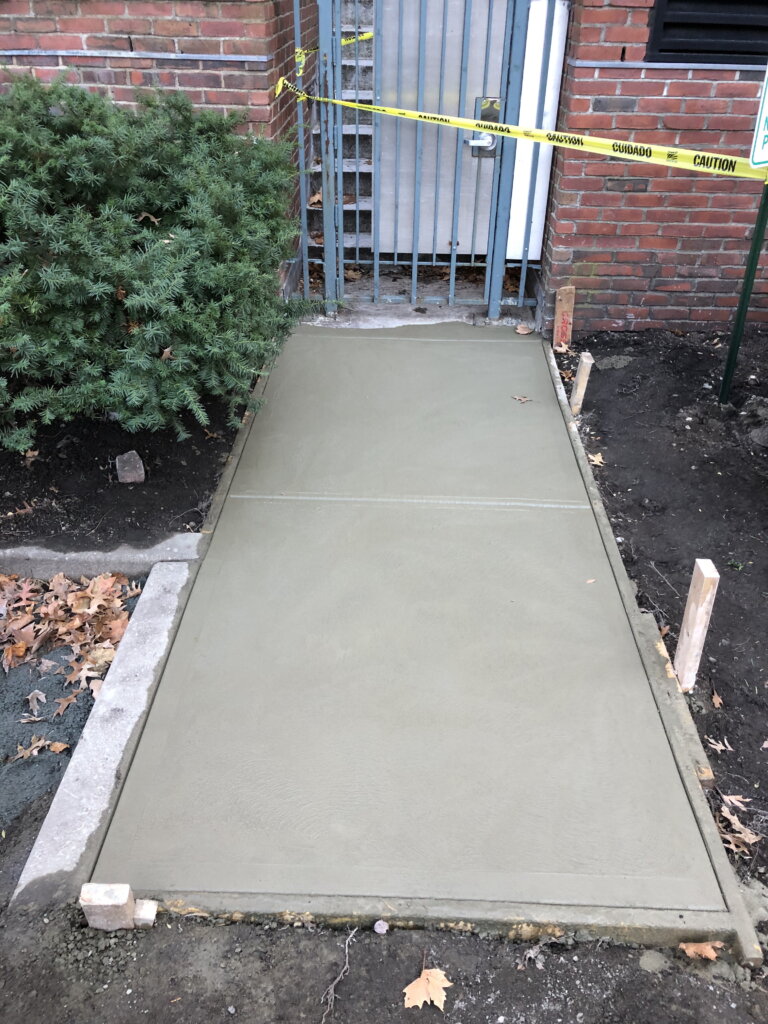 A freshly poured concrete walkway in front of a building, still wet and smooth, with wooden forms along the edges 