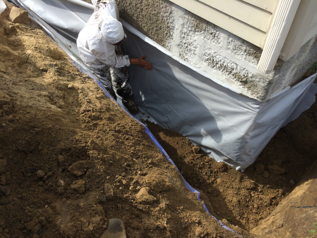 A worker standing beside a building foundation, applying a waterproofing wrap to the exterior concrete wall. 