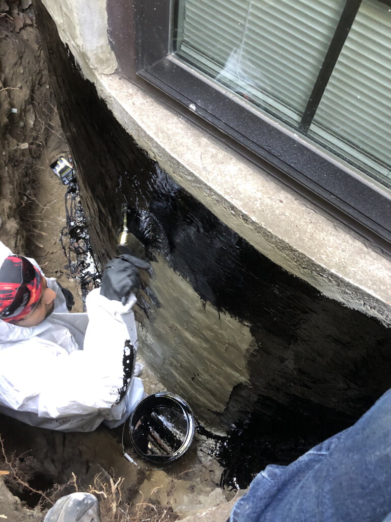 A person applying waterproofing material to the exterior of a building foundation.