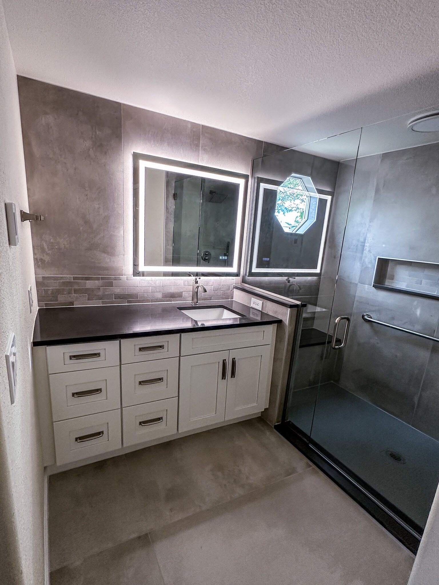 Finished primary bathroom with backlit vanity mirror, dark tile accent wall, black quartz countertop, and glass-enclosed walk-in shower