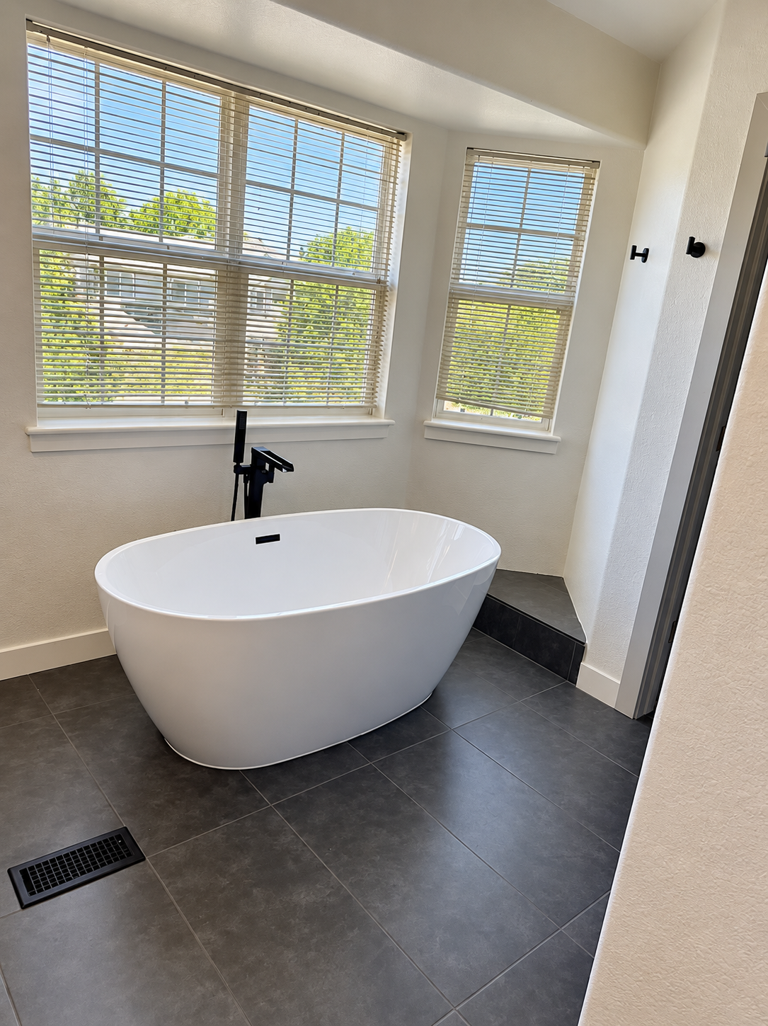Freestanding white tub beneath large windows with black floor-mount filler