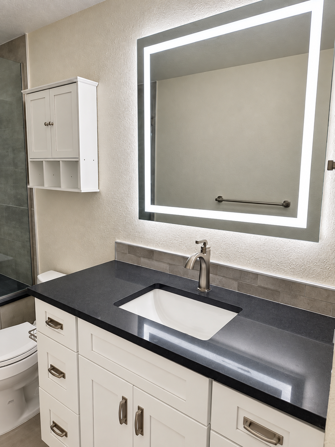 Bathroom vanity with dark countertop, white cabinetry, and backlit mirror