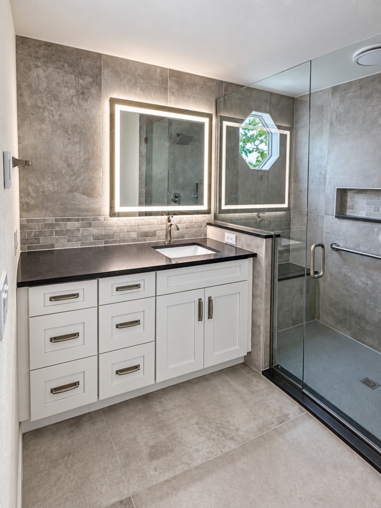 White bathroom vanity with gray tile and glass-enclosed shower, completed by The Build Squad.