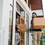 Two people carrying moving boxes out of a house
