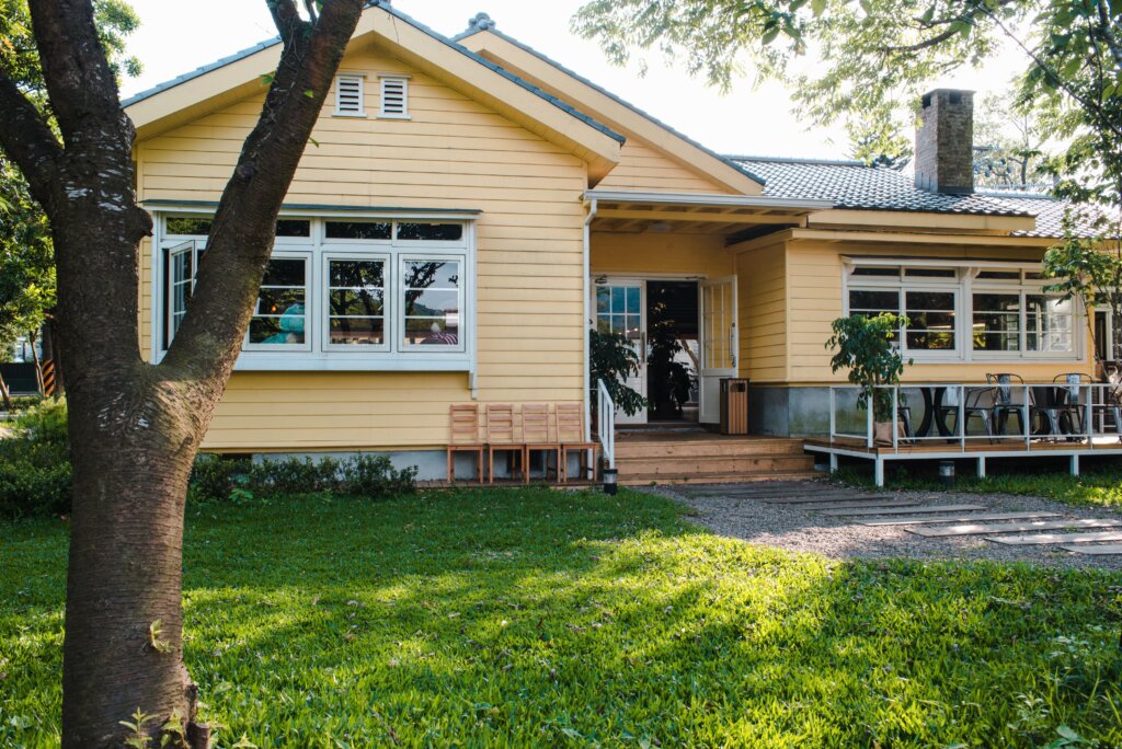 “Yellow single-story house with large windows, a covered porch, mature trees, and a spacious green lawn.”
