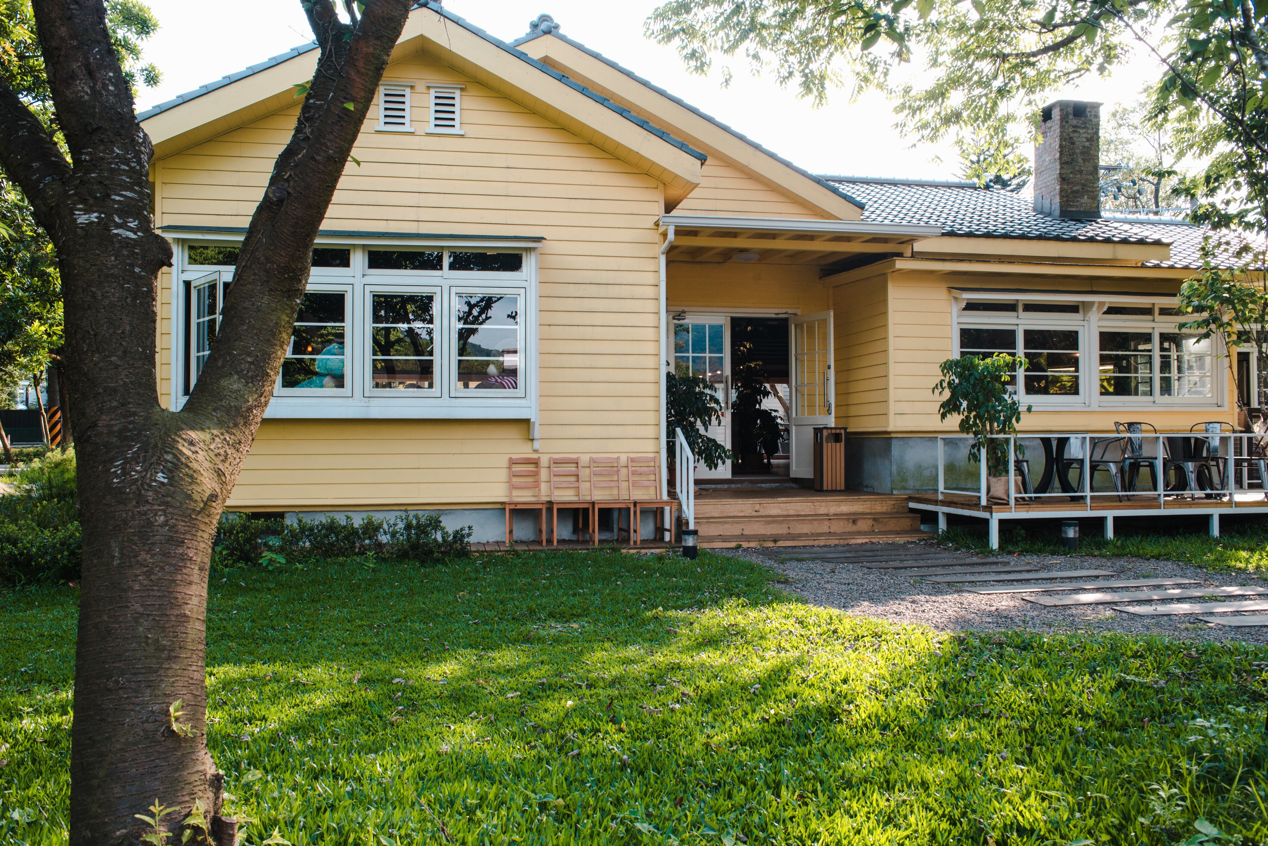 “Yellow single-story house with large windows, a covered porch, mature trees, and a spacious green lawn.”