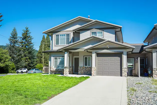 Modern two-story suburban house with a garage, green lawn, concrete driveway, and trees under a clear blue sky.