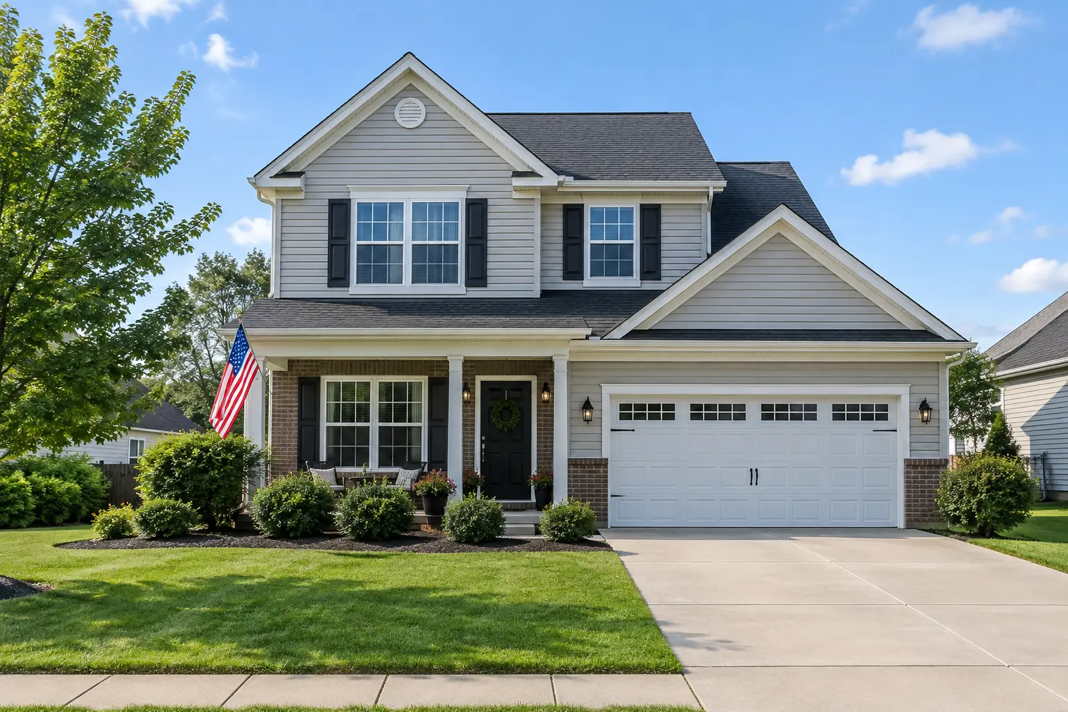 Gray suburban house with a white garage, front porch, landscaped lawn, and American flag.