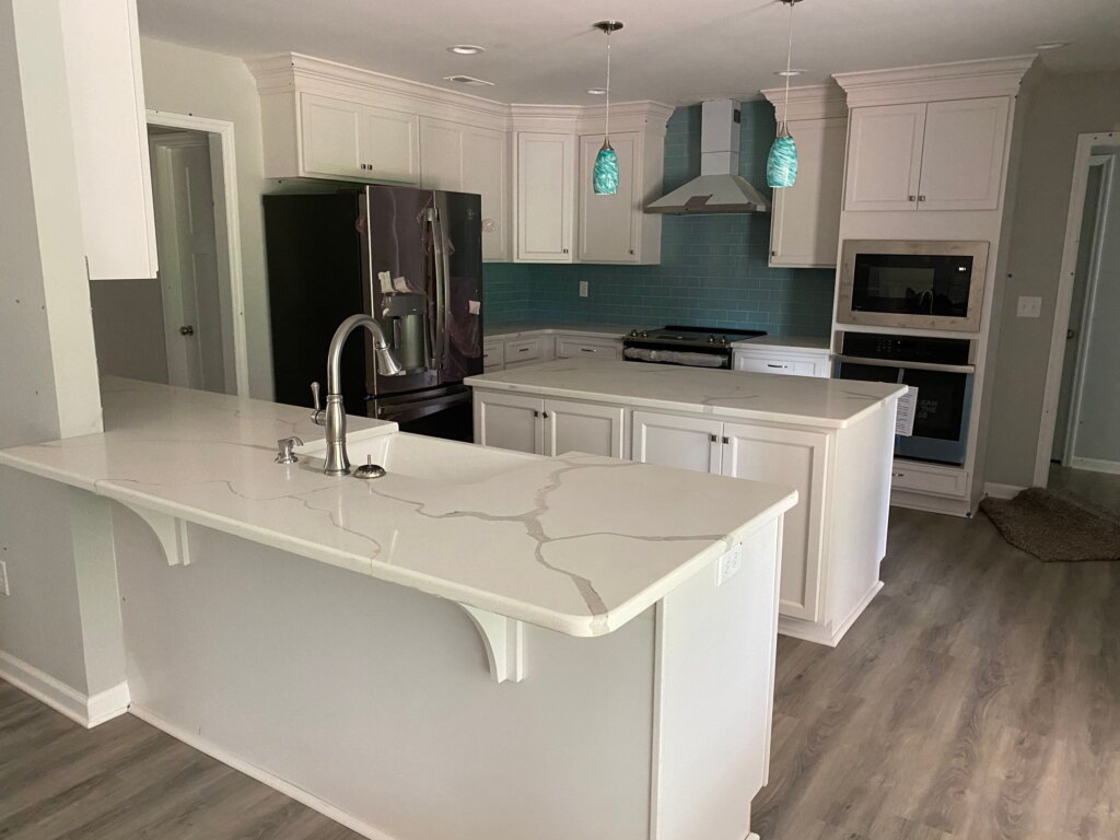 A bright, newly renovated kitchen featuring white cabinetry, a teal subway tile backsplash, white quartz countertops with grey veining, a central island, and a separate breakfast bar with a sink and gooseneck faucet.