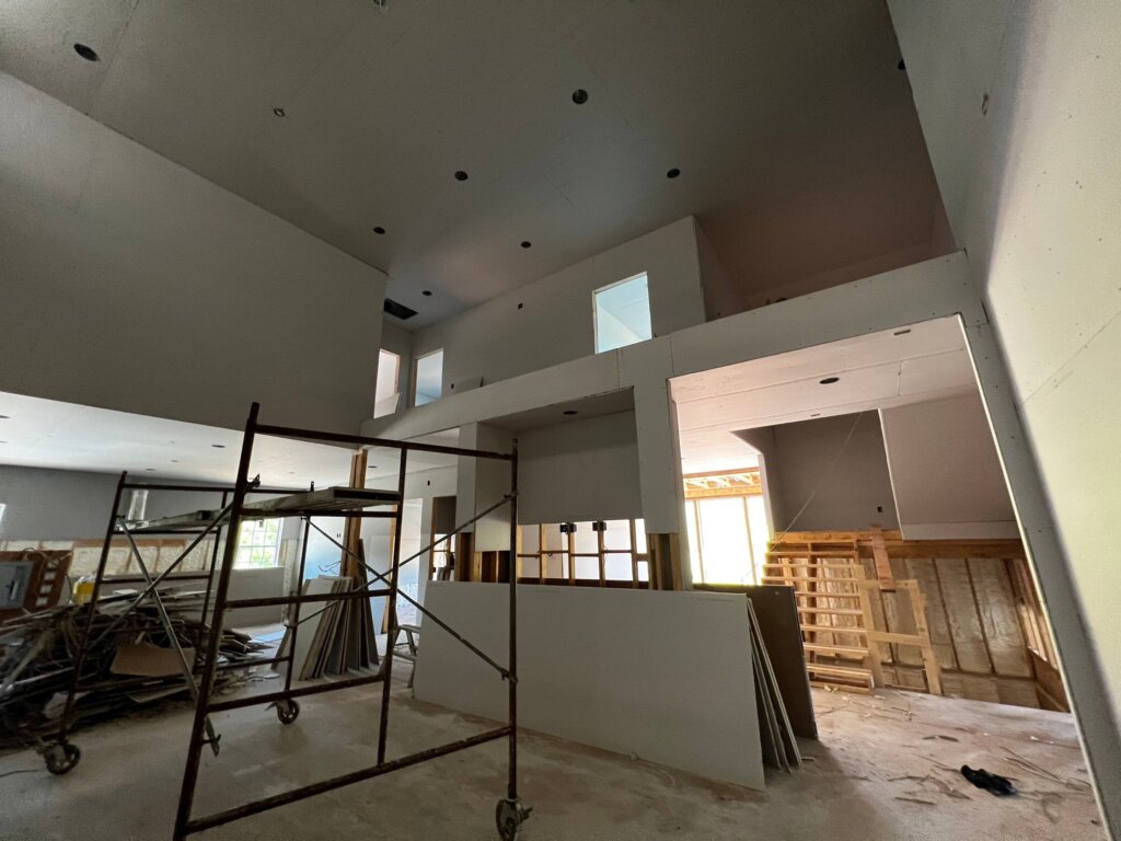 An interior view of a luxury custom home under construction with high ceilings, freshly hung drywall, scaffolding, and exposed wooden framing for a staircase.