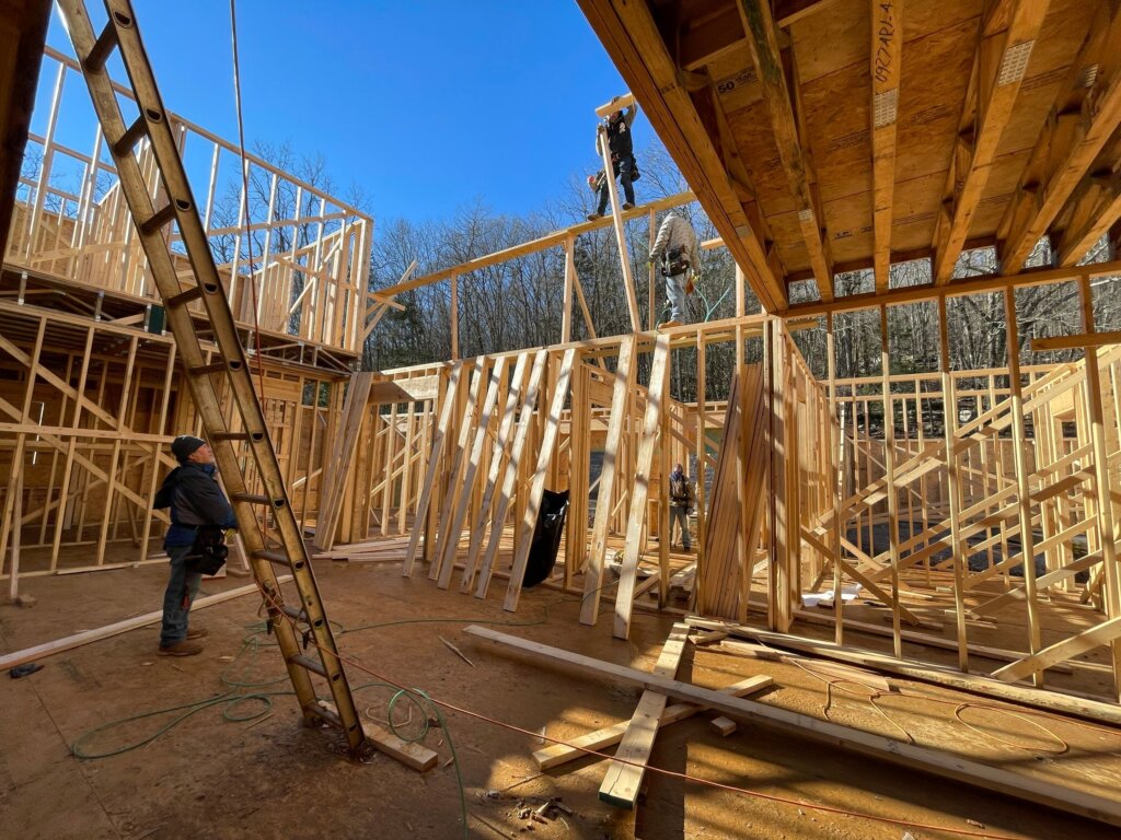 An open-air view of a multi-story custom home under construction, showing a network of exposed wooden wall studs and floor joists. Several construction workers are framing the structure under a bright blue sky.