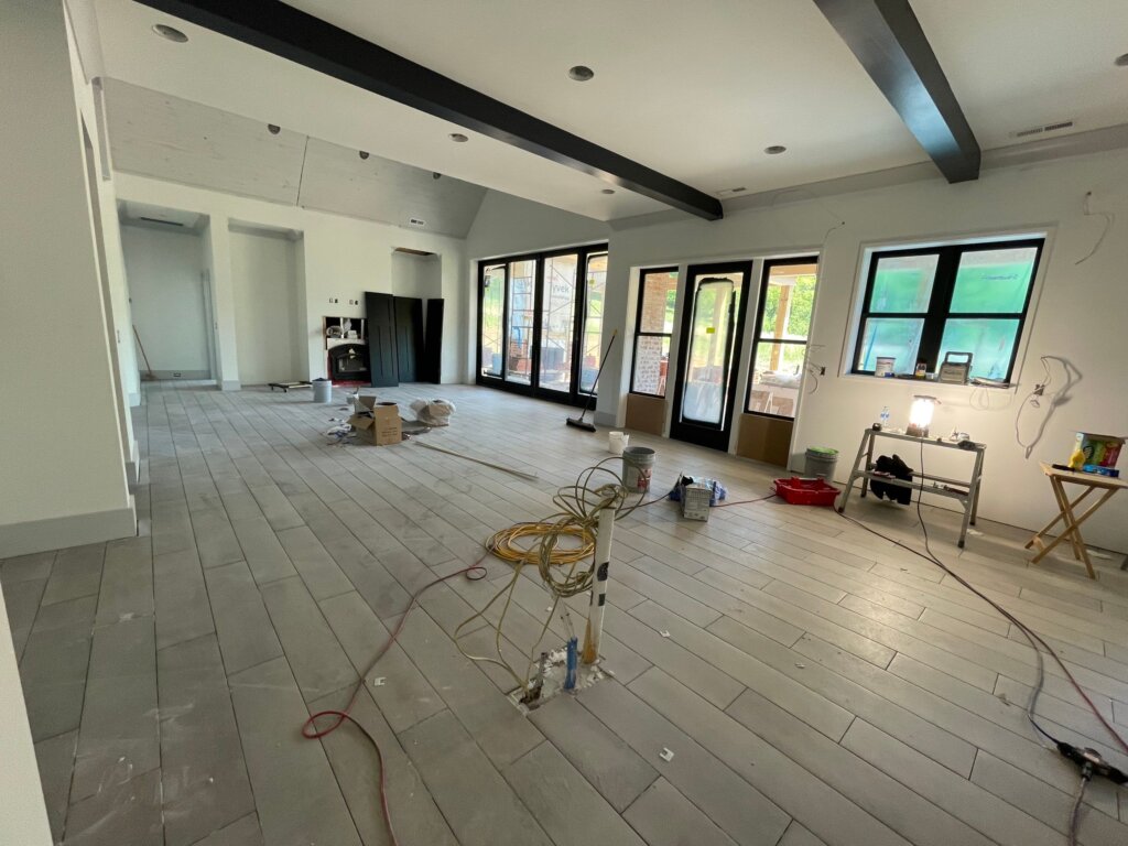 Open concept living space under construction featuring black exposed ceiling beams, light wood-look plank flooring, and large black-framed patio doors.