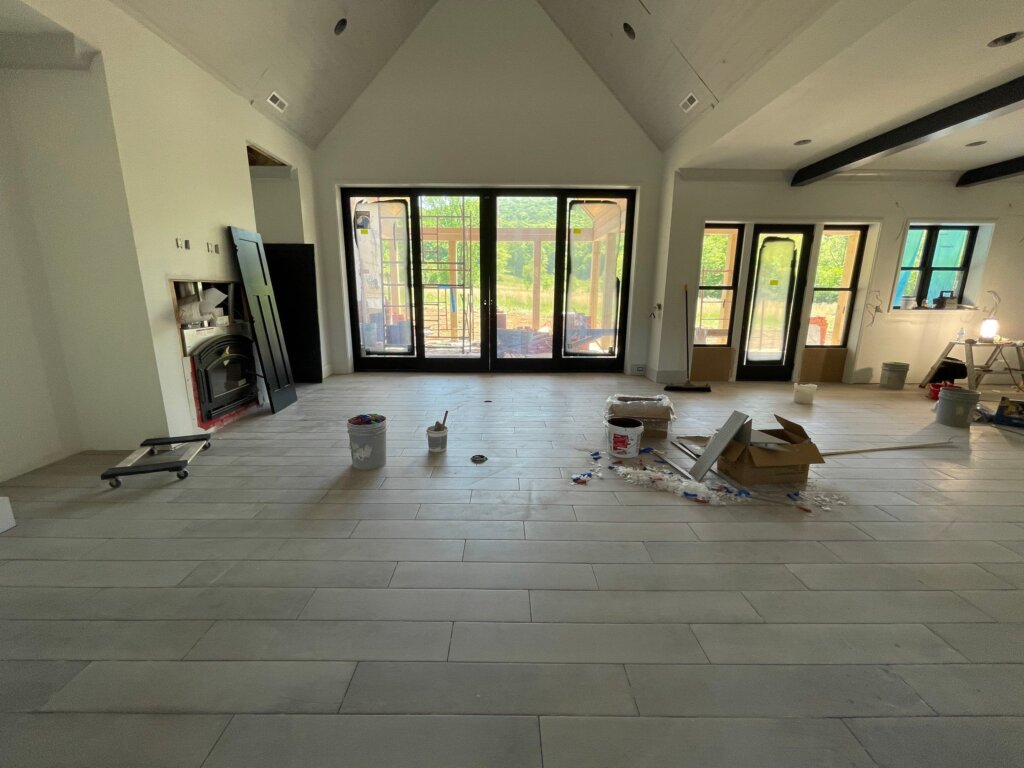 Large open-concept living room under construction featuring newly laid light gray wood-look plank tile flooring, a vaulted ceiling, and expansive black-trimmed sliding glass patio doors.