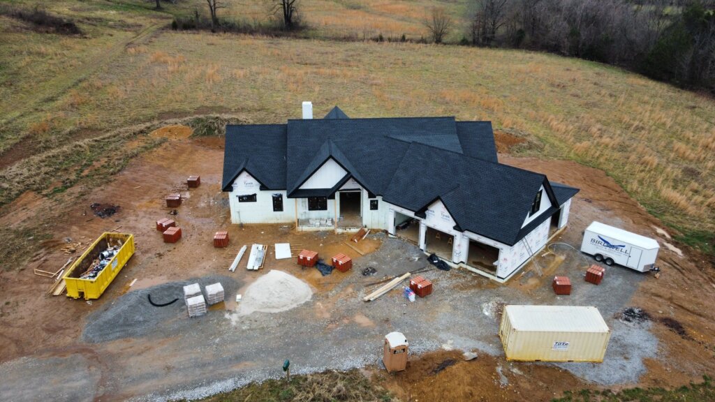 Aerial drone view of a modern farmhouse home construction site with a black shingle roof, white exterior walls, and a three-car garage.