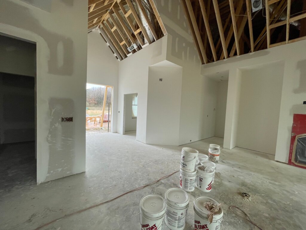 Custom home interior during the finishing stages, featuring floor tile installation, exposed subfloor electrical conduit wiring, and rolling drywall scaffolding visible outside.