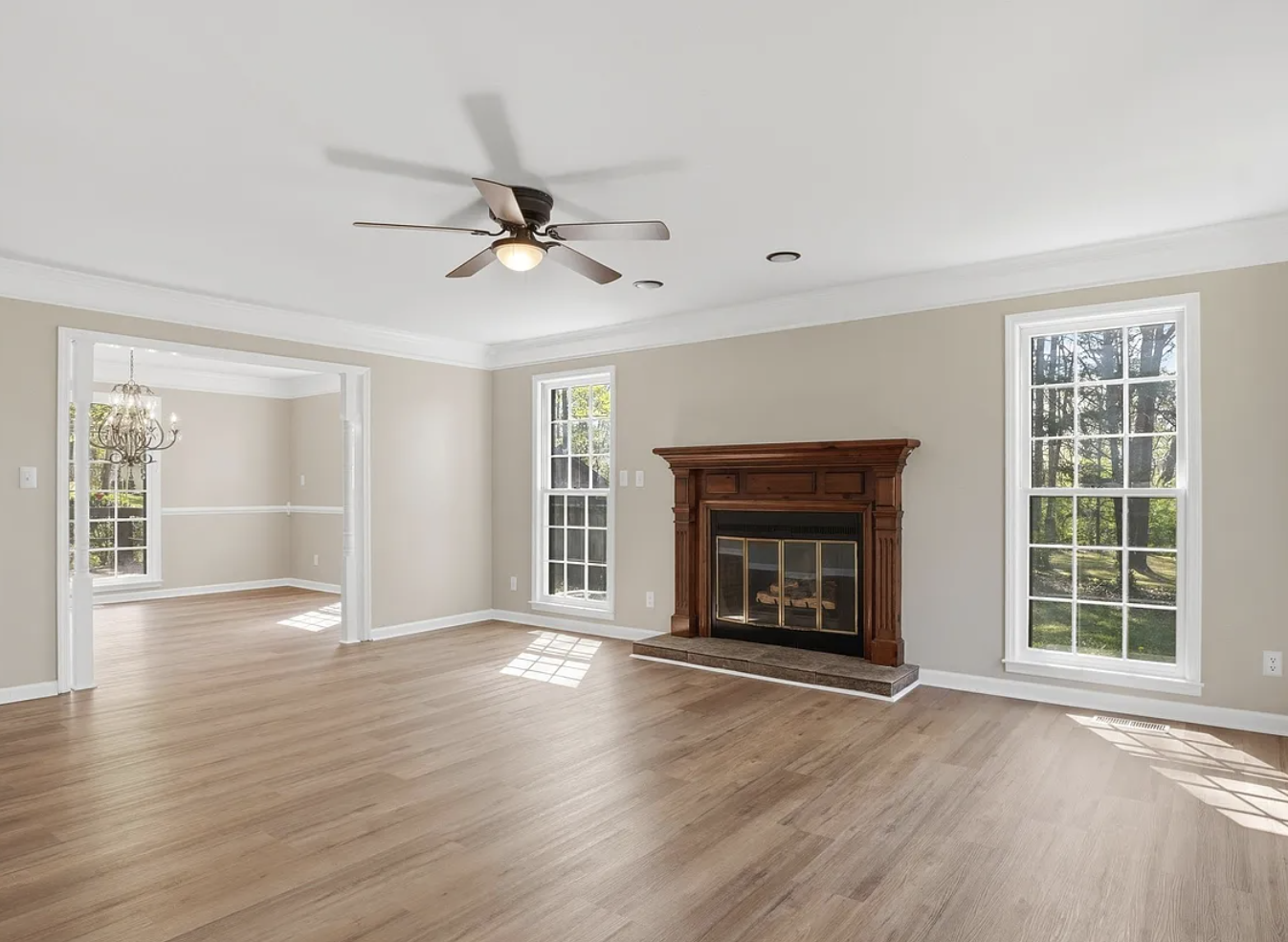 Completed living room renovation featuring light hardwood floors, a traditional stained wood fireplace mantel, beige walls with white crown molding, and a dark ceiling fan.