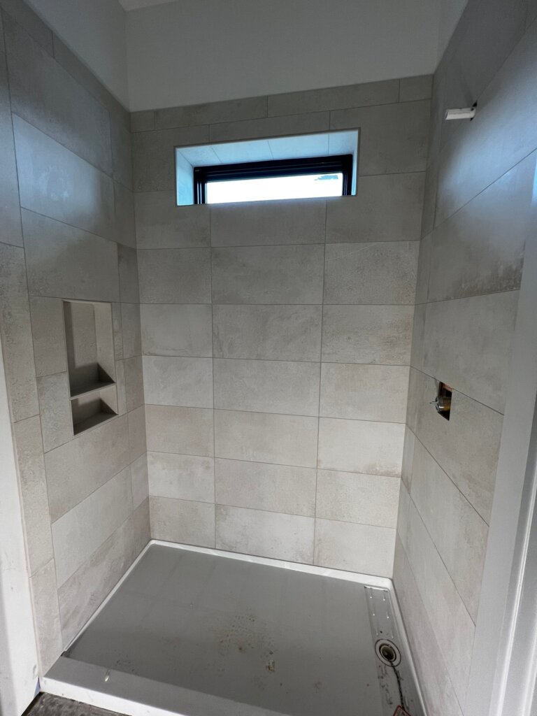A walk-in shower stall mid-construction, featuring large-format light grey wall tiles, a built-in recessed storage niche, a high rectangular transom window, and a white shower pan with exposed plumbing fixtures.