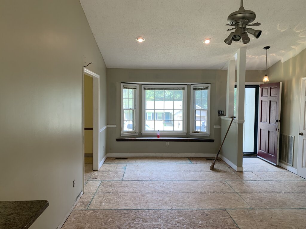 An empty living room or entryway mid-renovation, featuring exposed plywood subflooring, beige walls, a large bay window with a built-in seat, and an open maroon front door on the right.