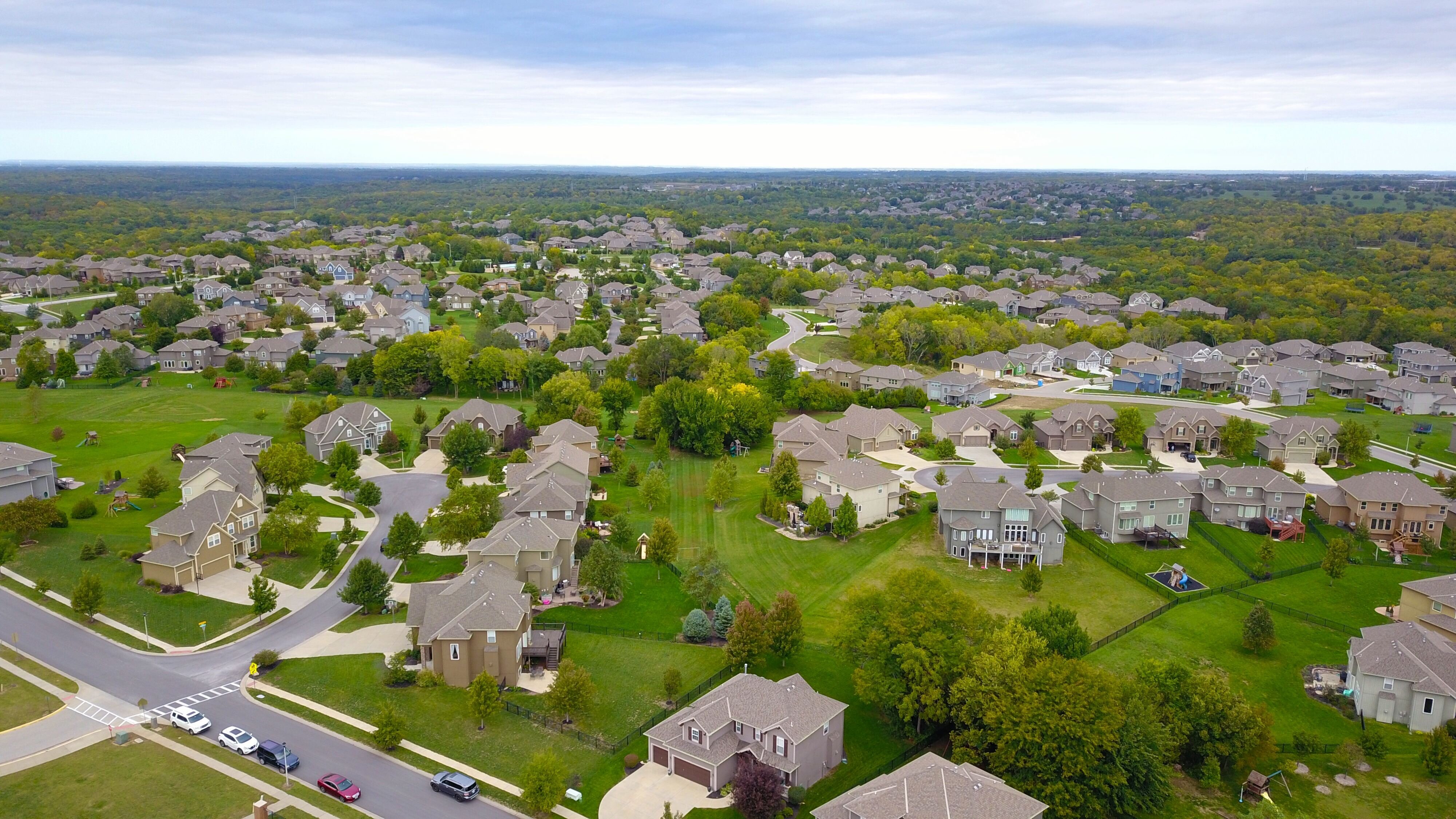 An aerial, high-angle view of a sprawling suburban neighborhood in North Alabama, featuring multi-story single-family homes with large green lawns, winding streets, and a dense treeline stretching into the horizon under an overcast sky.