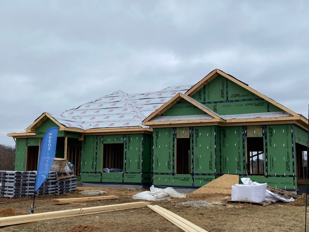Residential house framing in progress featuring exposed wood studs, roof joists, and weather-resistant wall panels on a concrete foundation slab.
